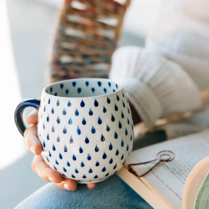 Woman holds a white ceramic coffee mug with blue raindrop pattern, while reading a book.